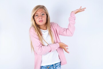 Fototapeta na wymiar caucasian blonde little girl wearing pink jacket and glasses over white wall pointing aside with both hands showing something strange and saying: I don't know what is this. Advertisement concept.