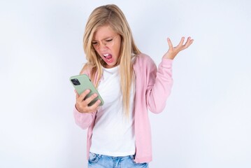 Photo of outraged annoyed caucasian blonde little girl wearing pink jacket and glasses over white baholds cell phone, makes call, argues with colleague,  expresses negative emotions. People and anger.