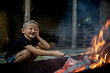 Fototapeta na wymiar The boy is sitting near the fire and burning sticks. The child spends time outdoors in the summer