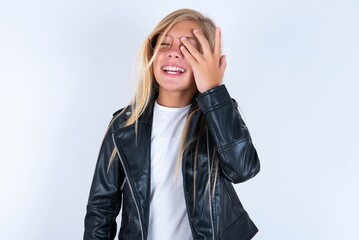 beautiful caucasian blonde little girl wearing biker jacket and glasses over white background makes face palm and smiles broadly, giggles positively hears funny joke poses