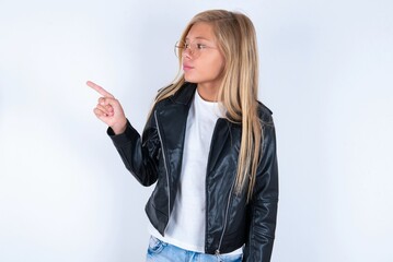 beautiful caucasian blonde little girl wearing biker jacket and glasses over white background points at copy space and advertises something, advices best price.