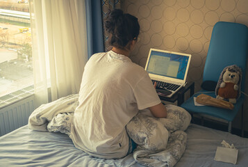 Teen girl sitting on the edge of the bed, using a laptop at home, girl chatting with friends on social networks, spending an interesting weekend, typing the text of a book, shopping online,