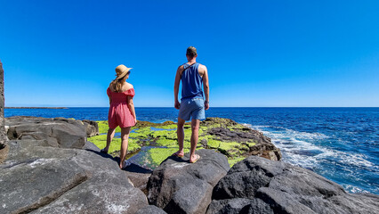 Couple with scenic view on volcanic rocks overgrown by moss, green sea plants, algae on coastline in Puerto de la Cruz, Tenerife, Canary Islands, Spain, Europe. Ocean bath Laja de la Sal. Wave hitting