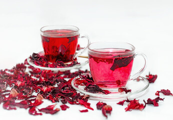 Two cups of Red hot hibiscus tea in a glass mug on a white table among dry Sudanese rose flowers.