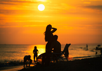 Dreamy evening landscape on beach with silhouettes of chilling on th sand people, hugging couple and children swimming in the sea during golden sunset