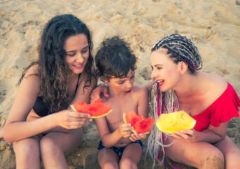 family, leisure and people concept - happy mother and two children having picnic on summer beach and eating red and yellow watermelon