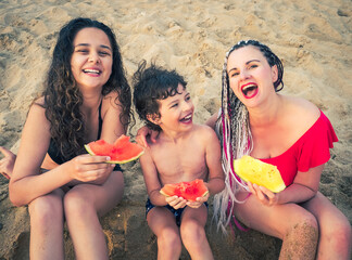 family, leisure and people concept - happy mother and two children having picnic on summer beach and eating red and yellow watermelon