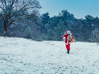 Happy Santa Claus with a big bag of gifts walks merrily to the side in a snowy winter forest. Christmas holiday concept.