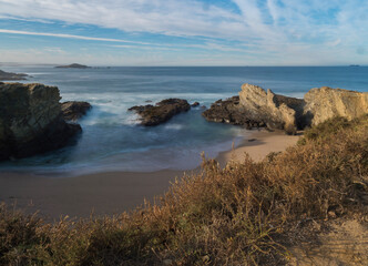 View of empty Praia dos Buizinhos sand beach with long exposure blurred ocean waves and sharp rock and cllifs at wild Vicentina coast in Porto Covo, Portugal. Golden hour.