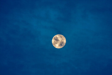 View of an illuminated full moon on a blue sky with dark clouds.