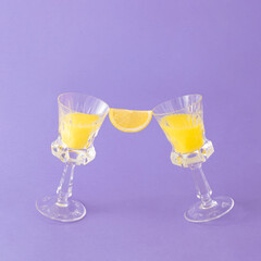 Lemon slice between two crystal glasses with yellow drink against purple background. Minimal cocktail party concept. Holidays celebrations aesthetic.