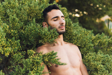 Muscular naked ethnic guy lying on green bush with closed eyes