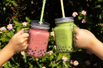 two unrecognisable girls toast with jugs with reeds and lids, milk tea and tapioca bubbles, against a background of green flowering plants,