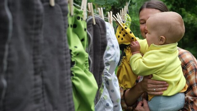 Mother with little baby exploring reusable diapers drying on clothesline in the garden. Modern eco friendly cloth nappy for infant child hygiene. Sustainable lifestyle, zero waste concept.