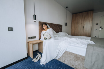 Obraz na płótnie Canvas A beautiful, young bride model is sitting on a bed in a bedroom, a hotel room in a white dress with a long veil with a bouquet of roses. Wedding portrait, photography.