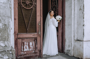 Obraz premium A beautiful, young bride in a white long dress stands near an old wooden door outdoors with a bouquet of roses in her hands. Wedding portrait, photography.