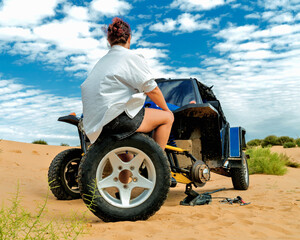 A young lady in shorts sits on a wheel taken from a buggy in the desert.
