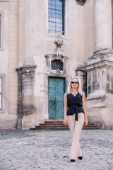 Fototapeta na wymiar A young, blonde girl in a black blouse and sunglasses poses against the backdrop of an old courtyard in Lviv. Ukraine.