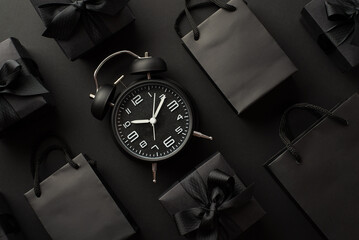 Black friday sales concept. Top view photo of black alarm clock gift boxes with ribbon bows and trendy paper bags on isolated black background - obrazy, fototapety, plakaty