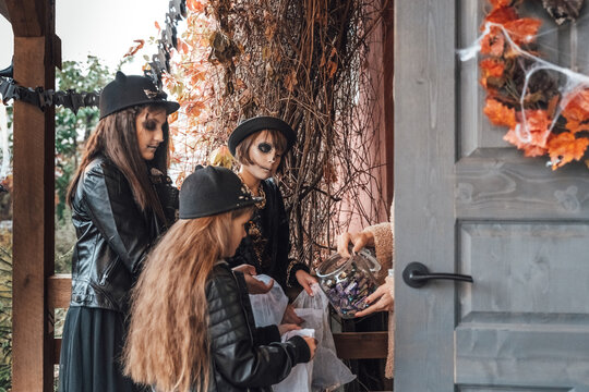 Scary girls, sisters, friends celebrating halloween. Playing treak or treat game on porch with garland. Bags with sweets in hands.Terrifying face skull makeup.Witch stylish costumes. Children's party