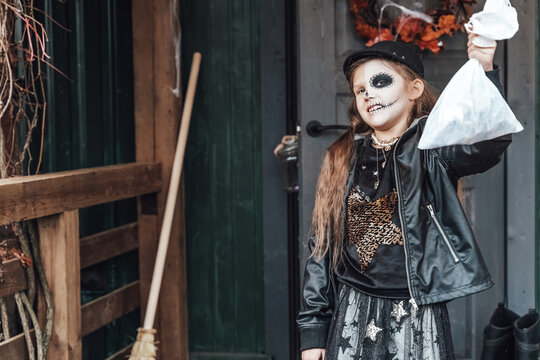 Scary girl celebrating halloween. Playing treak or treat game on porch with garland. Bag with sweets in hands.Terrifying face skull makeup.Witch stylish costumes. Children's party