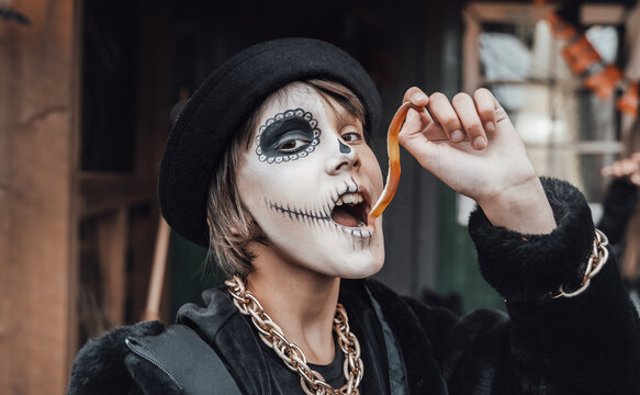 Beautiful scary little girl celebrating halloween, chewing gummy worm. Terrifying face makeup and witch costume, stylish image. Horror, fun at children's party in barn on street. Hat, fur coat, chain