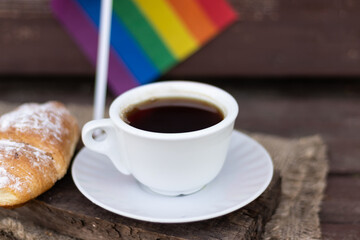 A cup of coffee and a croissant on a wooden table against the background of the LGBT flag