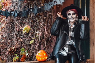 Scary woman,girl celebrating halloween.Terrifying black skull half-face skeleton makeup.Witch costume,stylish image. Jacket, hat.Fun at photoshoot,holiday party on autumn porch.Pumpkin jack-o-lantern