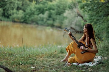 A hippie woman playing her guitar smiles and sings songs in nature sitting on a plaid in the...