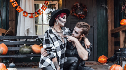 Fototapeta premium Scary love family couple man,woman celebrating halloween.Terrifying black skull half-face makeup,witch costumes.Stylish images,hat.Photoshoot,holiday party. Decorating of porch,pumpkin jack-o-lantern