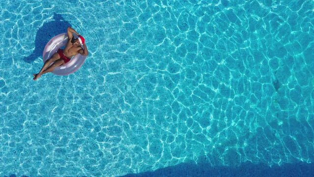 Drone shot of man on Christmas holiday floating in inflatable ring in swimming pool wearing Santa hat - shot in slow motion