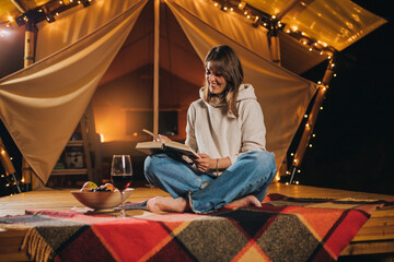 Obraz na płótnie Canvas Smiling Woman freelancer drinking wine and read book sitting in cozy glamping tent in autumn evening. Luxury camping tent for outdoor holiday and vacation. Lifestyle concept