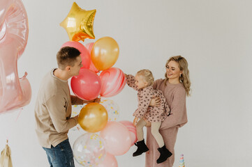 A little girl with mom and dad hug and kiss on the background of a cake and balloons. Cake smash