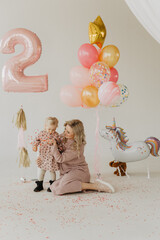 Mom with a little daughter in the studio against the background of balloons, confetti and a cake.