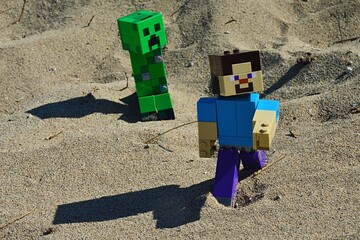 Obraz premium LEGO Minecraft figure of main character Steve chased by green explosive Creeper mob on sandy dunes, summer afternoon sunshine.