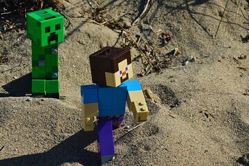 Obraz premium LEGO Minecraft large figure of Steve chased by hostile explosive green Creeper mob on arid sandy landscape in summer afternoon sunshine. 