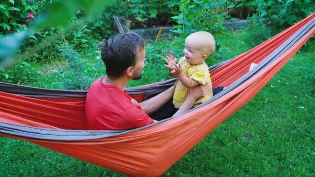 Father and his little toddler child daughter playing clapping hands in hammock in garden. Family resting on backyard at summer day.