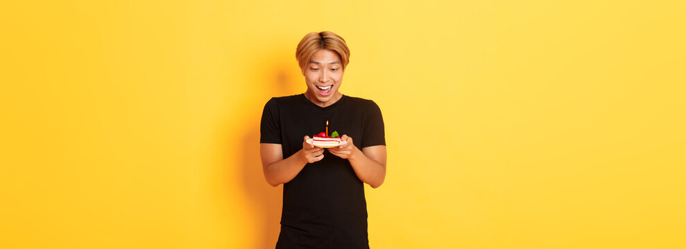 Excited Happy Attractive Asian Guy Smiling As Looking At Birthday Cake, Making Wish, Celebrating B-day, Standing Yellow Background