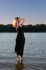 Young happy woman in white dress on shore of lake. One blond model on a beach during sunset. 