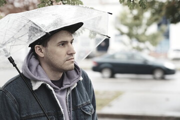 Young pensive man standing with transparent umbrella in rain on city street.