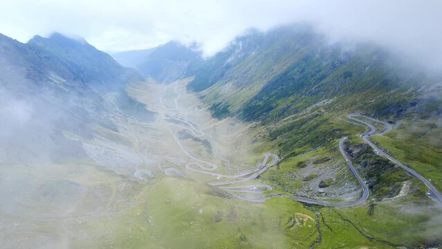 Aerial drone view of nature in Romania. Transfagarasan route in Carpathian mountains with moving cars visible through clouds, rocky slopes with greenery