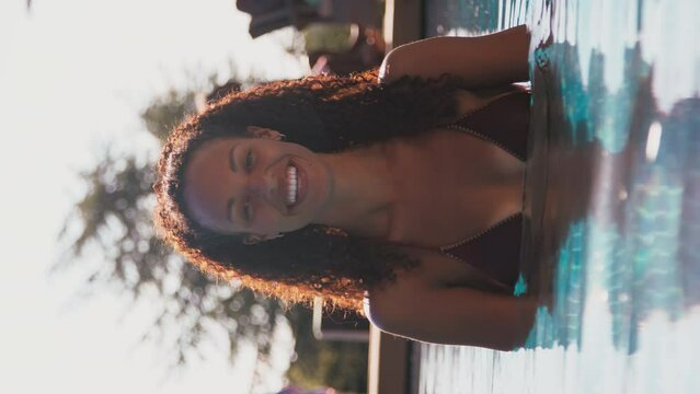 Vertical video of woman on summer holiday or vacation standing at the edge of swimming pool and smiling into camera - shot in slow motion