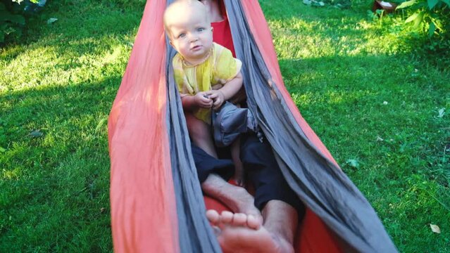 Curious cute little toddler child daughter sitting with her father in hammock. Family resting at backyard garden in summer sunny day.