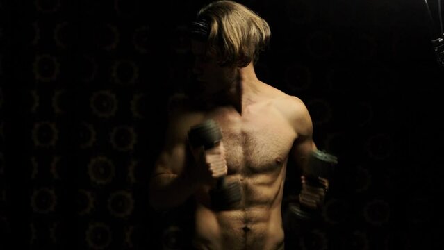 lean muscular man shakes his biceps with dumbbells in the dark at home.