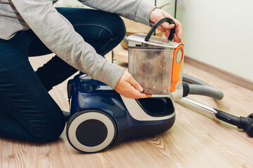 Woman takes container with dust filter out of canister vacuum cleaner at home. Container full of...