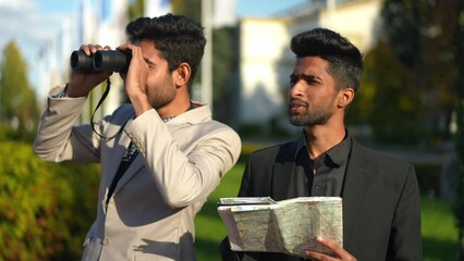 Two elegant Middle Eastern young men standing on urban city street using paper map and binoculars. Positive smiling handsome friends travelling talking outdoors. Business trip and leisure concept - Powered by Adobe