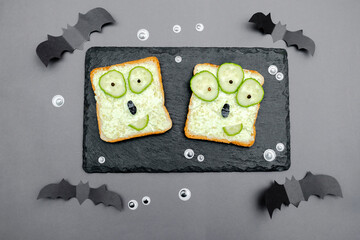 Funny monster faces on halloween sandwich toast bread with soft green avocado cheese ricotta butter,cucumbers on cutting serving board,bats on gray background.Kids child breakfast lunch food.