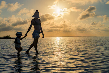 Fototapeta na wymiar Mother and son holding hands and walking in the water at sunset