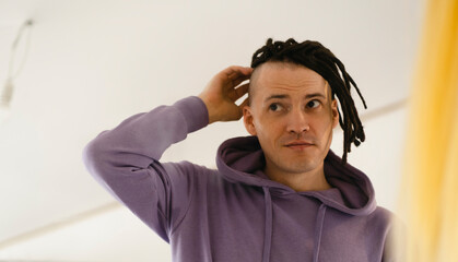 Young man looking in mirror, considering his new dreadlocks. Handsome guy preening in front of mirror in hairdressing salon.