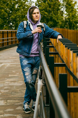 Young handsome man with dreadlocks in casual clothes drinking coffee from paper cup standing on footbridge in city park.